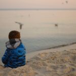 A boy sitting on the sand looking at sea